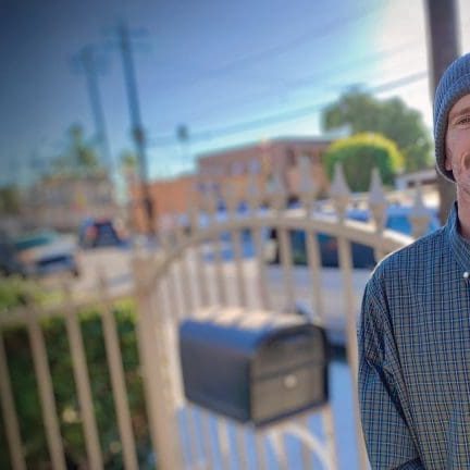 A man in a plaid shirt and beanie stands in front of a metal fence, with an urban street scene blurred in the background, embodying the vibe of 2024's end-of-year giving spirit.