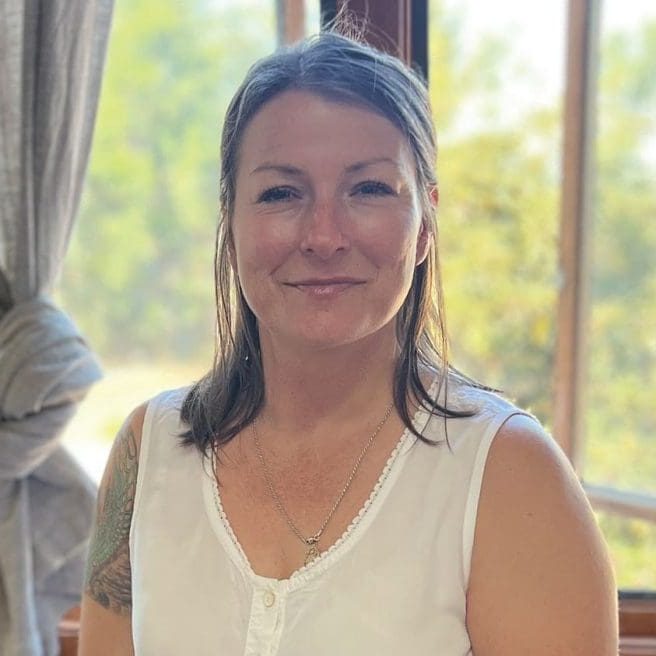 A woman with shoulder-length hair smiles indoors by a window, the vibrant trees outside symbolizing connection and community.