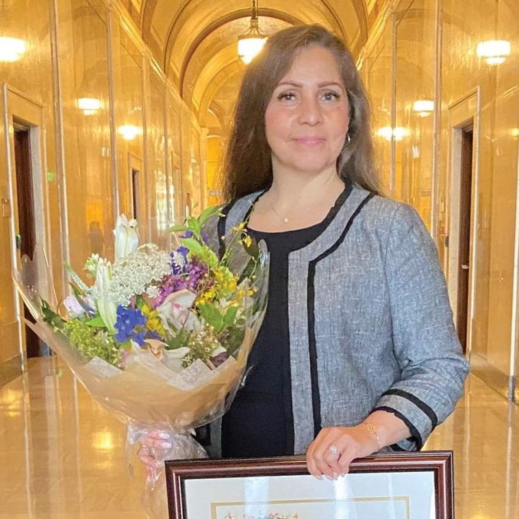 Josie Casarrubias stands in a hallway of the RMA San Fernando Valley, holding a bouquet of flowers and a framed certificate. She wears a gray jacket over a black top, embodying her role as Director with pride and elegance.