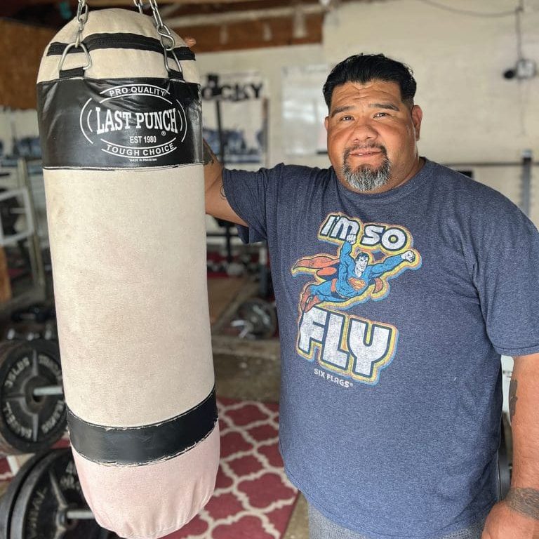 A man stands next to a punching bag in his home gym, wearing a blue Im So Fly Superman-themed T-shirt, showcasing the strength grace can bestow on ones program of fitness and determination.